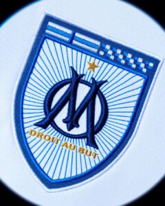 SOUHtpLmlZFodnv | UAE SPORT STORE Embroidered shield patch with blue rays, overlapping 'AM' initials, gold star, and the motto 'DROIT AU BUT' in gold.