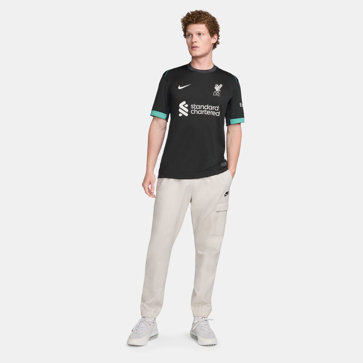 Full-body shot of a man wearing a black Liverpool FC training jersey with green trim and beige cargo pants