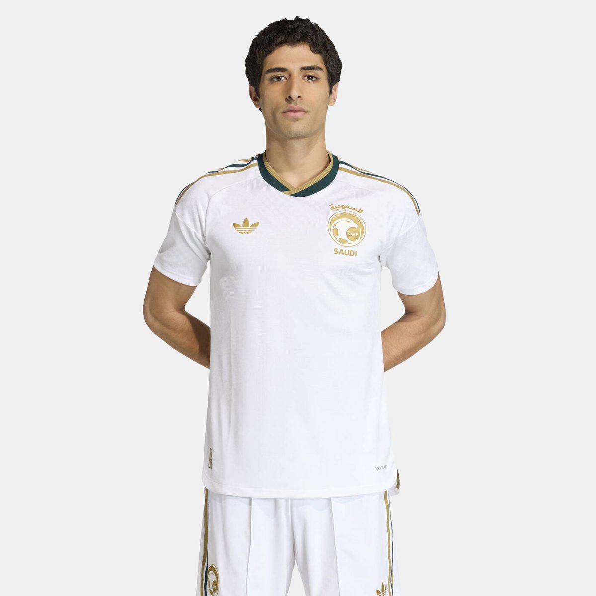 Male model wearing a white Saudi football jersey with a gold crest and Adidas logo, standing with hands behind his back.