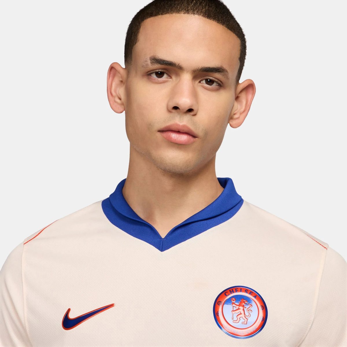 Close-up of a young man’s face and upper torso wearing a cream Chelsea FC football jersey with a blue collar; Nike swoosh and Chelsea crest are visible on the chest.