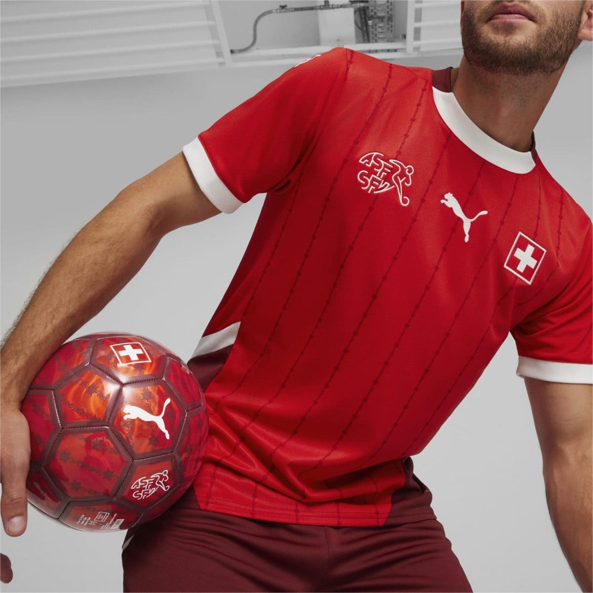 Soccer player in a red Swiss jersey holding a red patterned ball against a neutral gray background, showing Swiss and Puma logos on the shirt and ball.