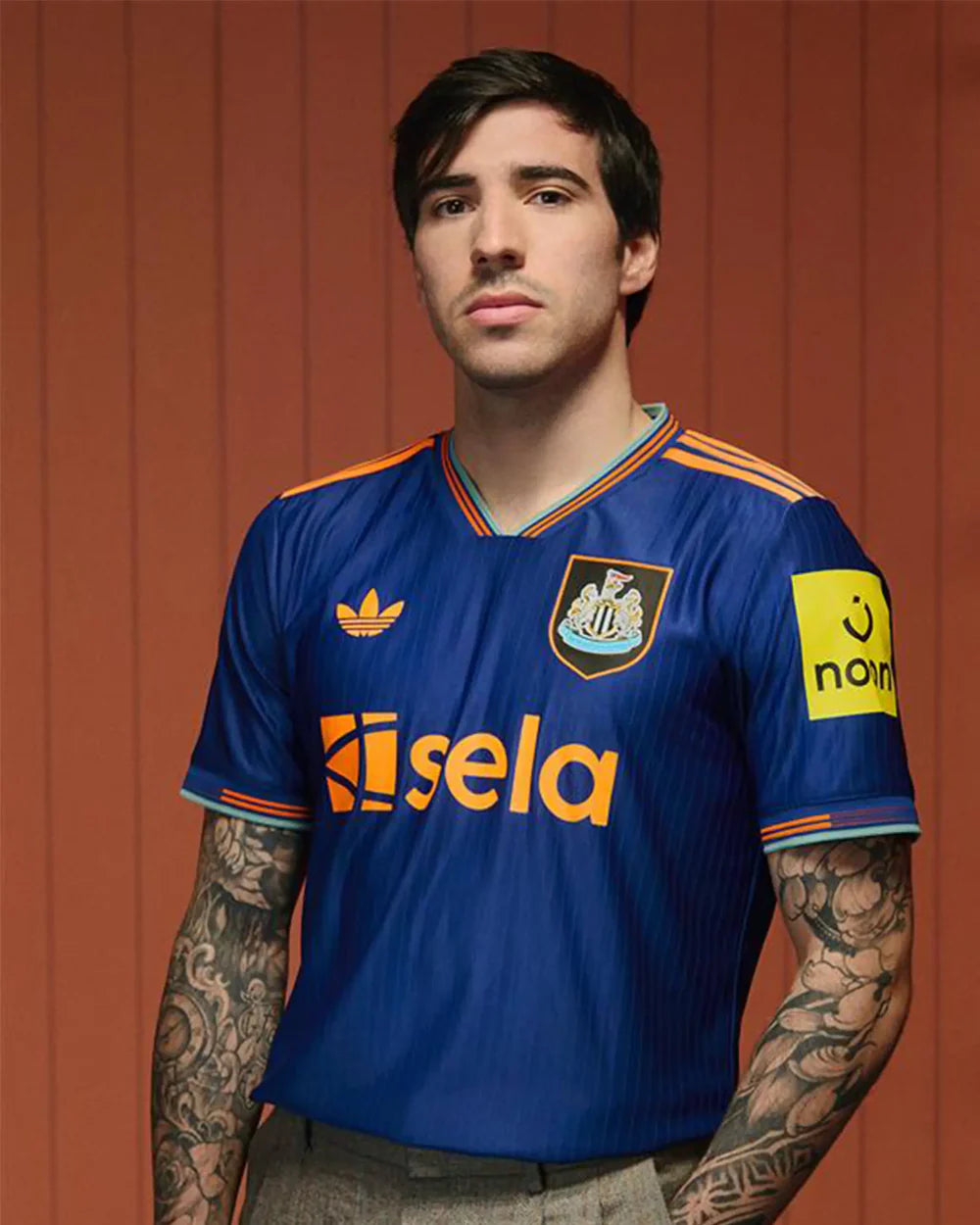Sandrothd-RESIZE | UAE SPORT STORE Portrait of a young man wearing a blue Newcastle United-style football jersey with orange trim, standing against an orange wooden backdrop.