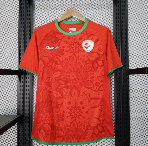 Orange soccer jersey with green trim on collar and sleeves, hanging on a wooden hanger; Kappa logo on the left chest and a crest on the right chest against a subtle floral pattern.