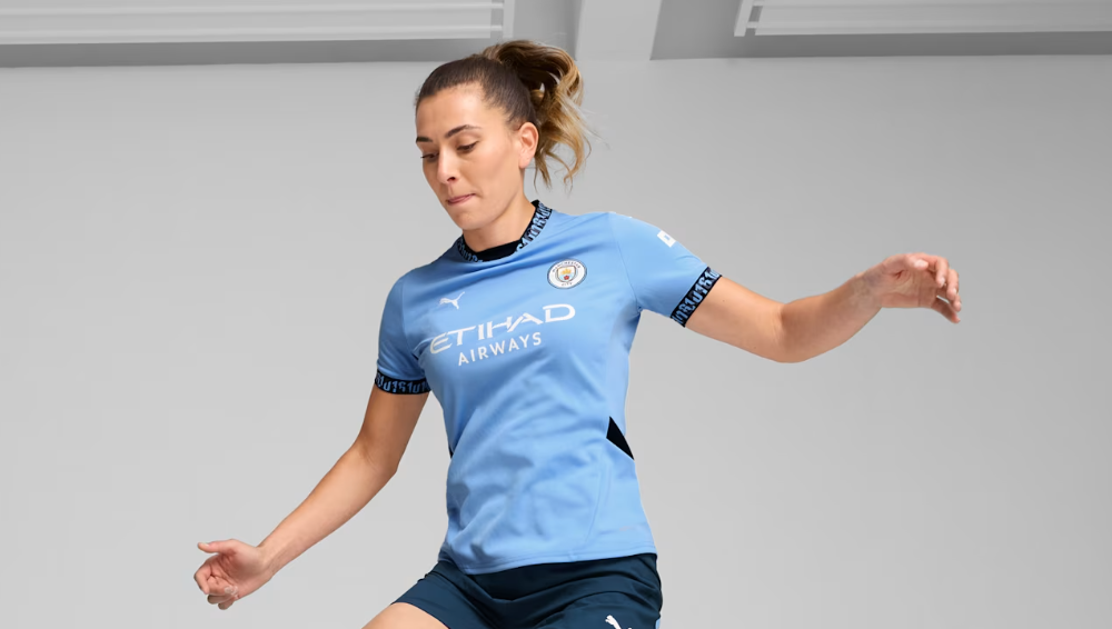 Female footballer in a light blue Manchester City kit mid-action, arms extended as she moves on the pitch.