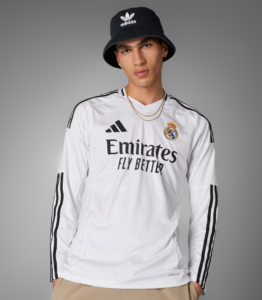 Male model wearing a Real Madrid white jersey with Emirates Fly Better logo, black Adidas bucket hat, and gold chains, posing against a gray backdrop.