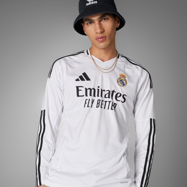 Male model wearing a Real Madrid white jersey with Emirates Fly Better logo, black Adidas bucket hat, and gold chains, posing against a gray backdrop.