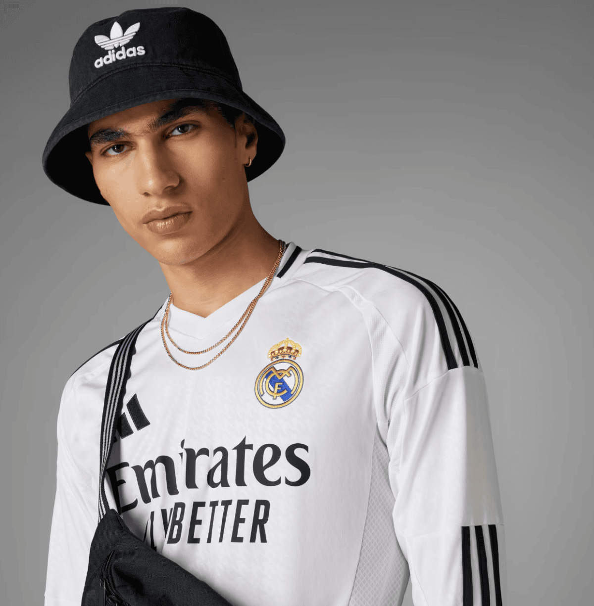 Portrait of a young man in a white Real Madrid jersey with Emirates branding, wearing a black Adidas bucket hat and a gold chain against a gray background.