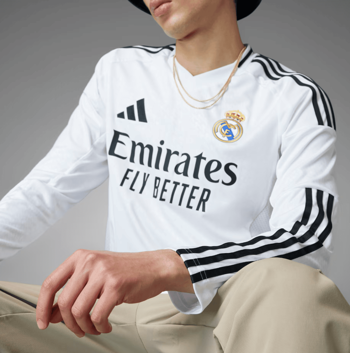 Person in a white Real Madrid jersey with Emirates Fly Better branding, wearing a black hat and layered gold chains, seated against a gray backdrop.