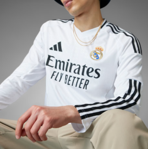Person in a white Real Madrid jersey with Emirates Fly Better branding, wearing a black hat and layered gold chains, seated against a gray backdrop.