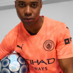 Close-up of a soccer player in a coral Manchester City jersey holding a blue and white patterned soccer ball.