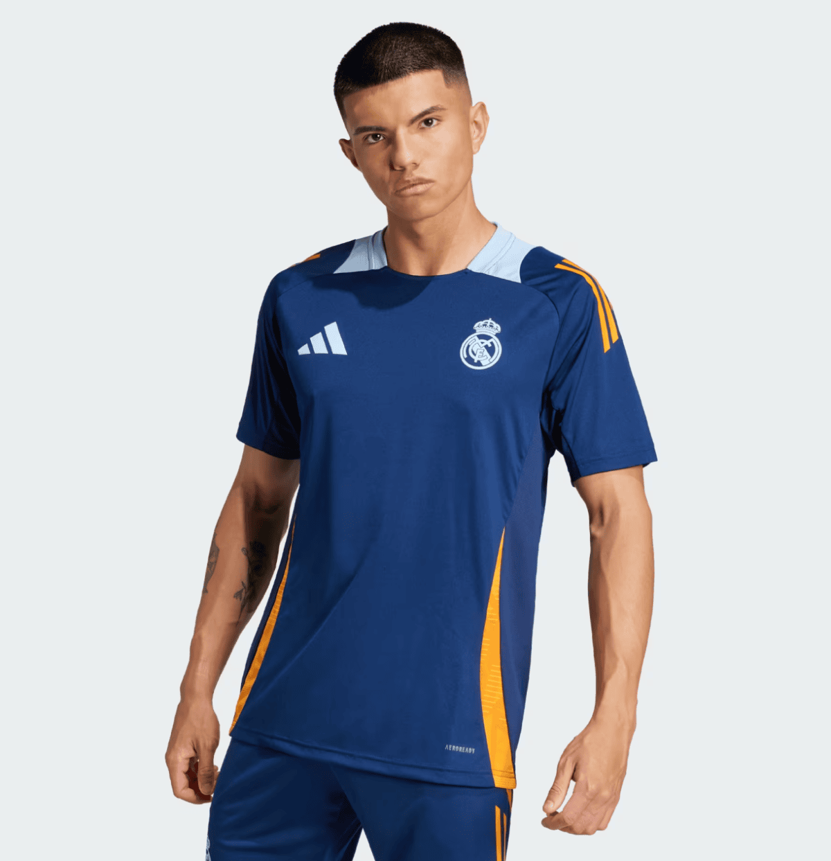 Male model wearing a blue Real Madrid away jersey with white collar and orange side panels, Adidas logo and Real Madrid crest visible against a light background.