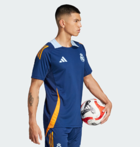 Male soccer player in blue Real Madrid kit holding a soccer ball at his side, profile view against a light background.