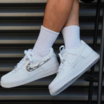 Pair of white Nike sneakers with a chrome wing design on the side, worn with white crew socks, resting on metal steps.
