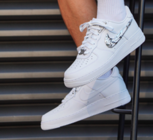 White Air Force 1 sneakers with a reflective chrome swoosh resting on metal stairs.