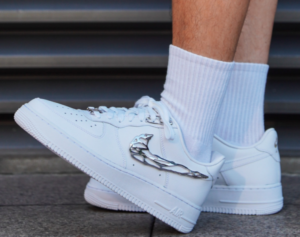 White sneakers with a metallic wing logo and white socks on a sidewalk in front of a metal grate. (informative)