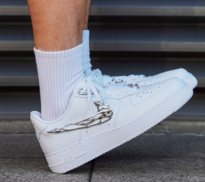 White Nike Air Force 1 sneaker with a silver chain detail around the heel, worn with a white sock against a dark background.