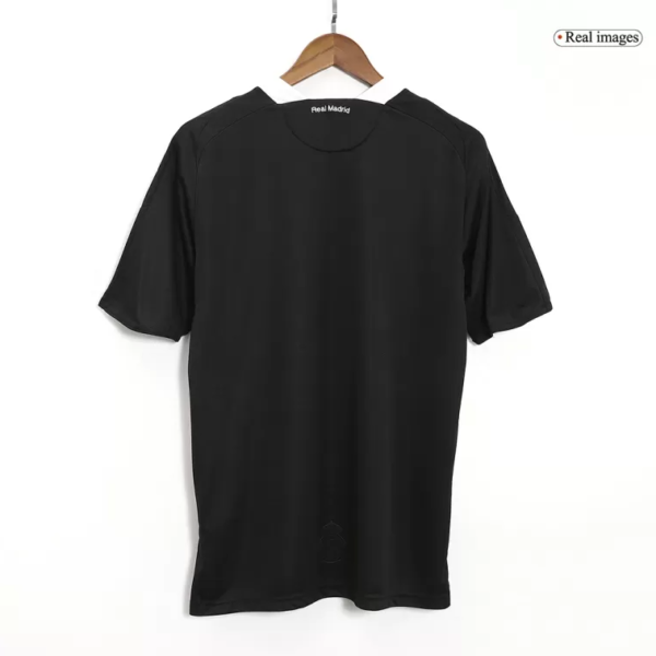Black Real Madrid jersey hanging on a wooden hanger, shown from the back with a small Real Madrid label at the neckline.