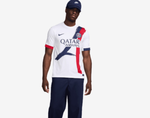 Male model wearing a white Paris Saint‑Germain jersey with Qatar Airways sponsor, navy/red brushstroke design, and navy cap.