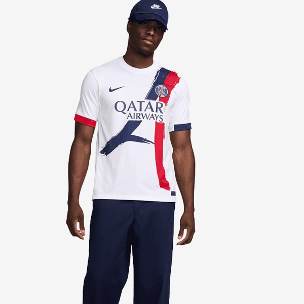 Male model wearing a white Paris Saint‑Germain jersey with Qatar Airways sponsor, navy/red brushstroke design, and navy cap.