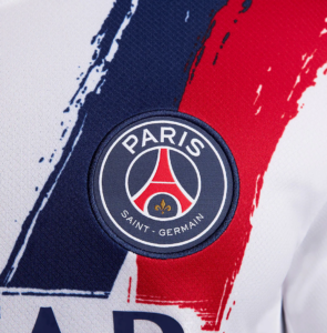 Close-up of the PSG badge sewn on a white jersey with blue and red brush strokes in the background of the kit.