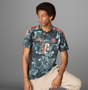 Man wearing a teal Bayern Munich soccer jersey with orange logos and beige pants, posing against a gray background.