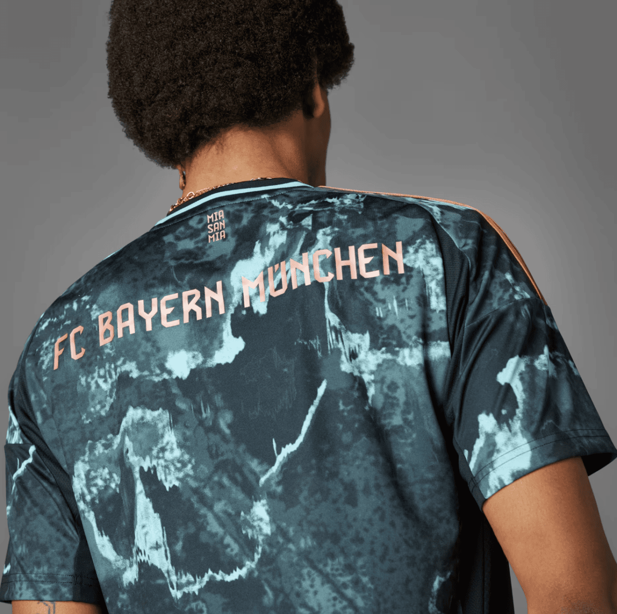 Back view of a person wearing a blue-green tie-dye FC Bayern München jersey with 'MIA SAN MIA' small text near the neckline.