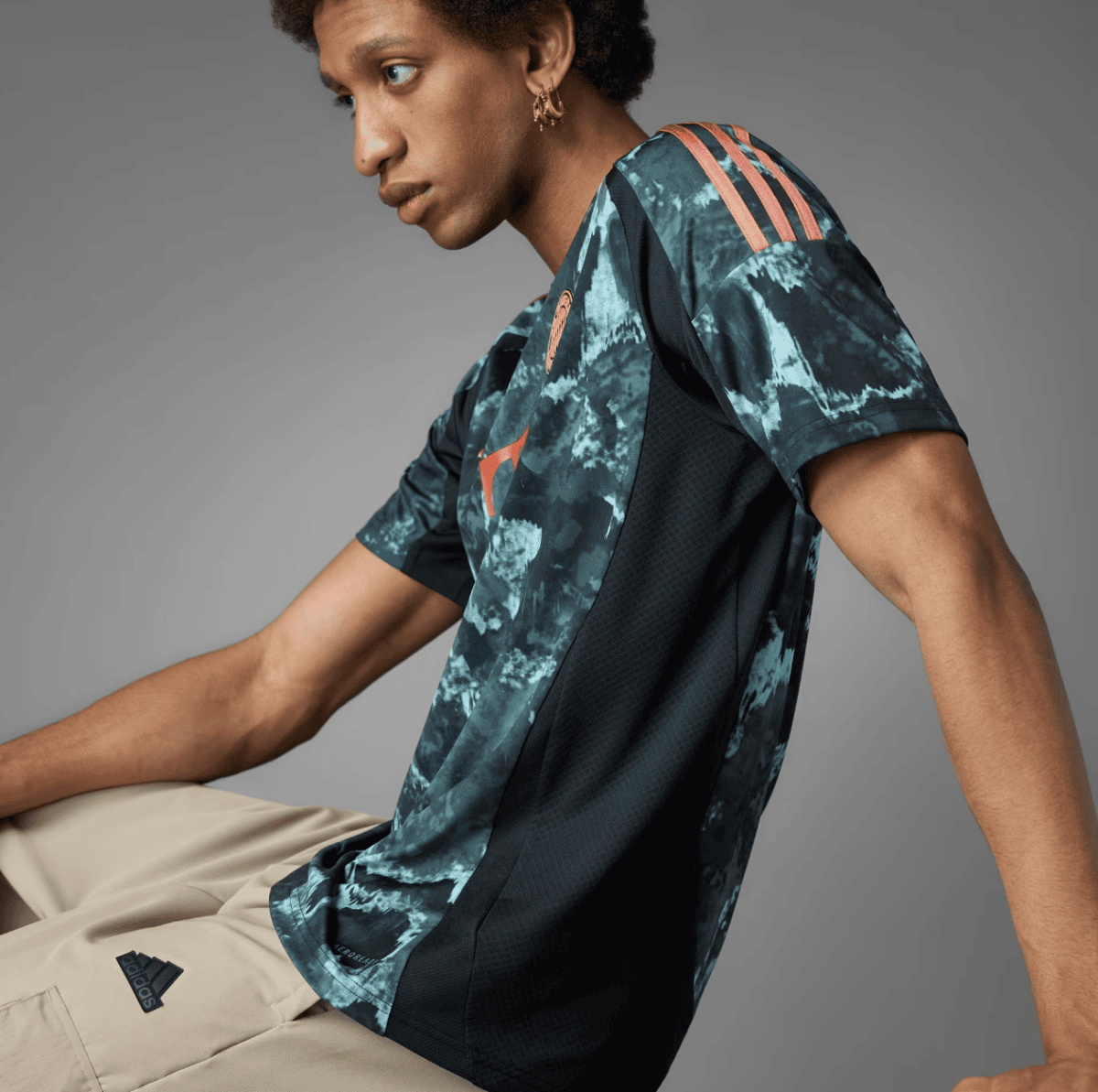 Side-profile of a young man wearing a teal and black tie-dye Adidas athletic shirt with orange logo, beige pants, and a gold hoop earring.