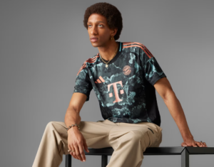 Young man seated on a black bench wearing a dark, marble-pattern Bayern Munich jersey with beige pants, against a gray backdrop.