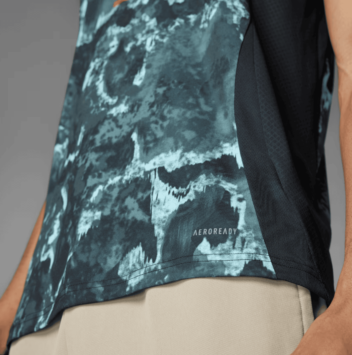 Close-up of a teal camouflage-pattern athletic shirt with a black side panel and AEROREADY logo, worn with beige shorts.