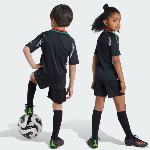 Two children in black soccer kits with green and red trim stand back-to-back; a boy rests his foot on a white soccer ball while a girl looks over her shoulder.