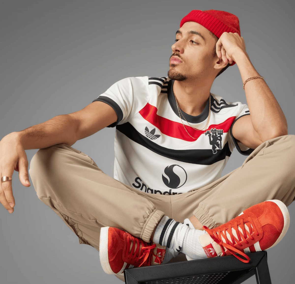 Young man in a red beanie sits cross‑legged on a chair, wearing a striped t‑shirt, beige pants, and red sneakers, looking off to the side on a gray background.