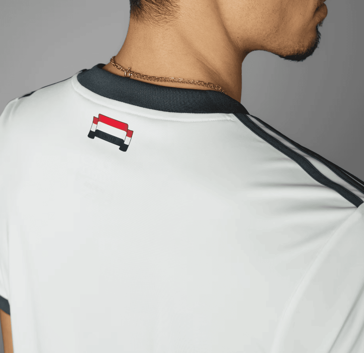 Back view of a person wearing a white sports shirt with a small Manchester United patch near the collar, and a gold chain necklace visible at the neck.