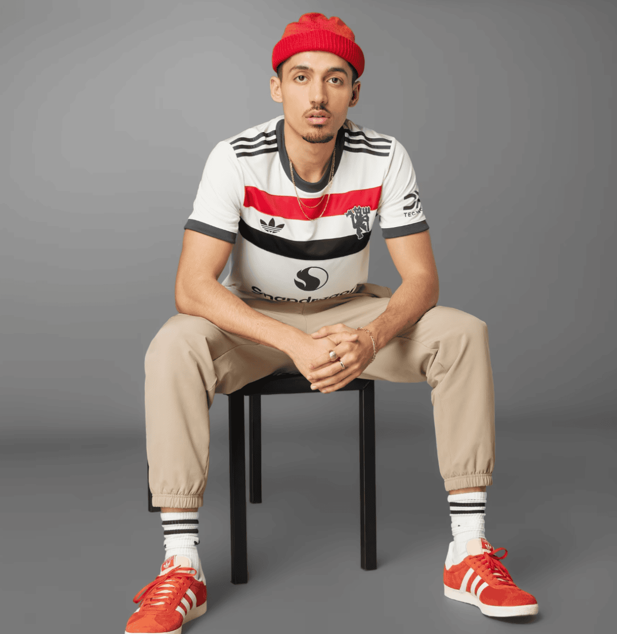 Young man sitting on a black stool wearing a red beanie, white striped t-shirt, beige joggers, white socks, and orange sneakers; hands clasped.