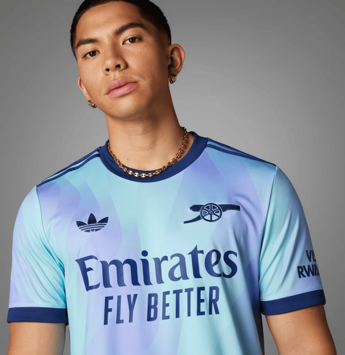 Portrait of a young man wearing a light blue gradient football jersey with the words 'Emirates Fly Better', plus a gold chain and hoop earrings, against a gray background.