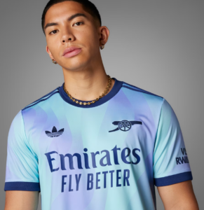 Screenshot_2024-09-18_194347_e9dc364f-38da-4e2e-be9b-9bf67388a332 | UAE SPORT STORE Portrait of a young man wearing a light blue gradient football jersey with the words 'Emirates Fly Better', plus a gold chain and hoop earrings, against a gray background.