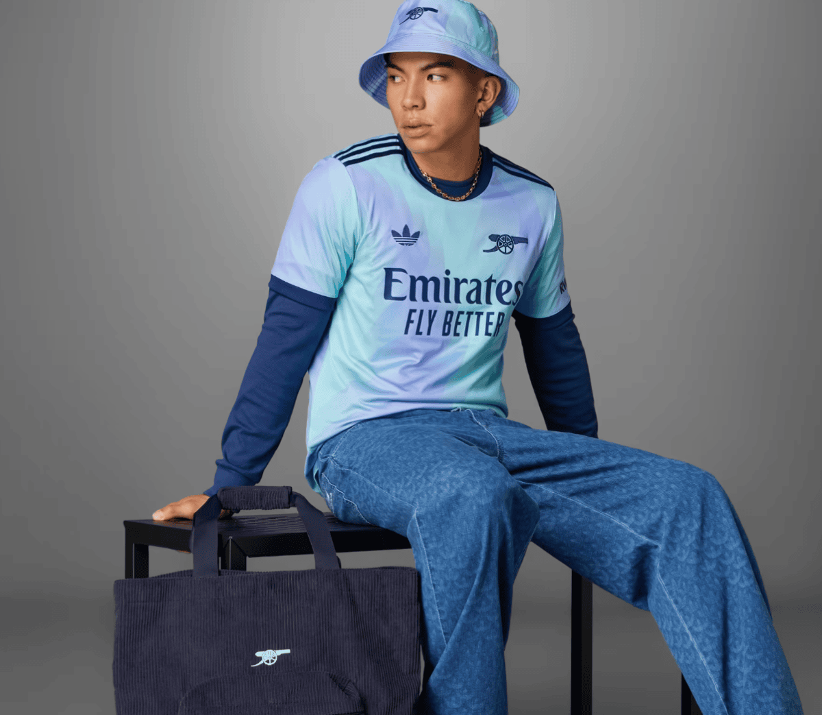 Person in a pastel blue Arsenal jersey and bucket hat sits on a black stool with a navy tote bag beside them.
