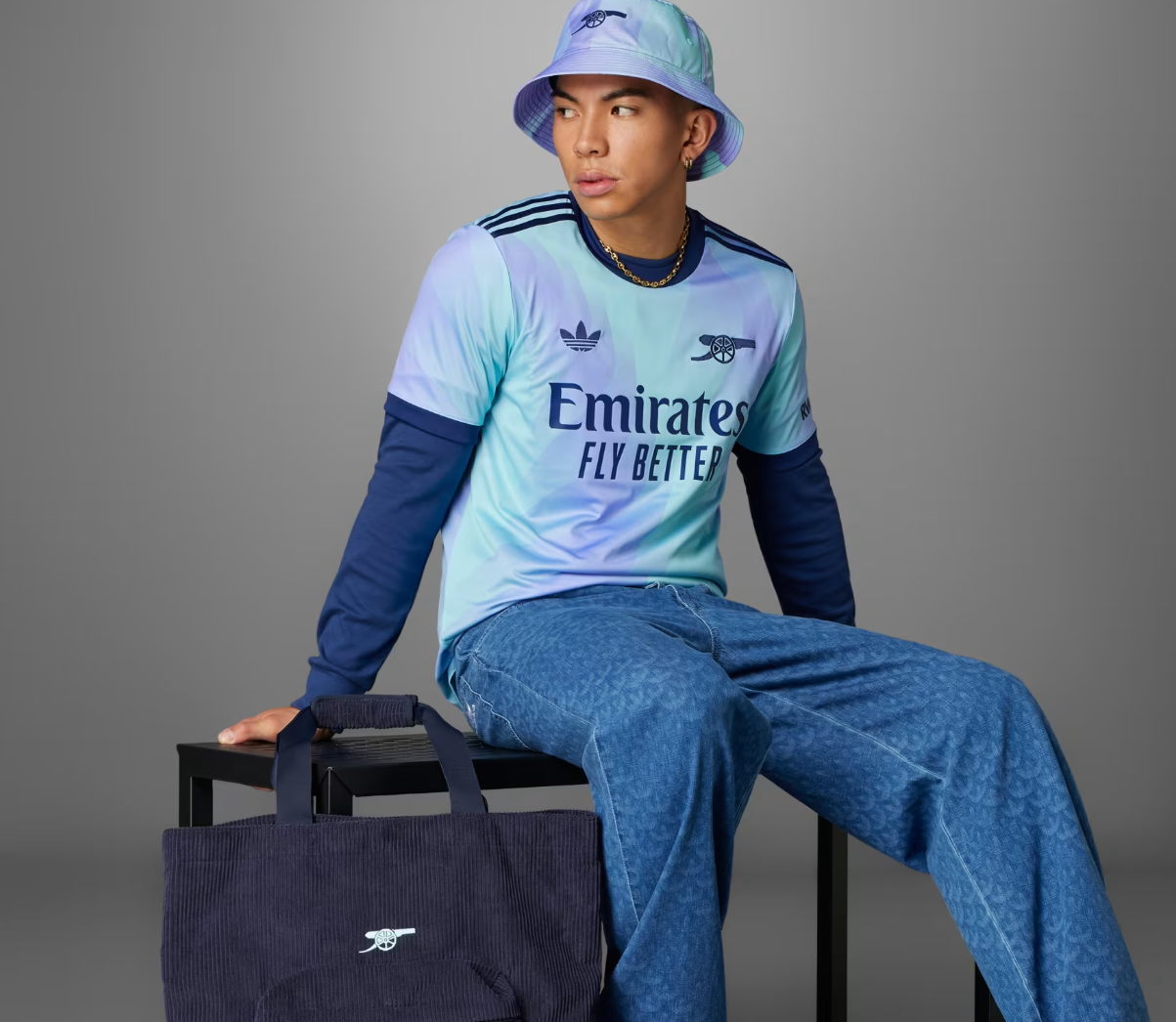 Screenshot_2024-09-18_194416_afaf008d-62d6-46a0-b159-846a3fd22383 | UAE SPORT STORE Person in a pastel blue Arsenal jersey and bucket hat sits on a black stool with a navy tote bag beside them.