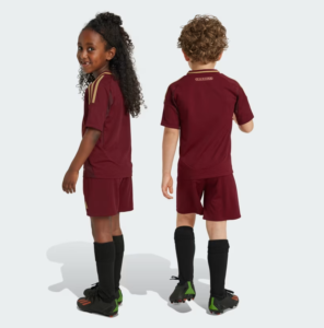 Two children in maroon soccer uniforms stand back-to-back, showing matching jerseys, shorts, long socks, and cleats against a light background.