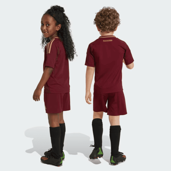 Two children in maroon soccer uniforms stand back-to-back, showing matching jerseys, shorts, long socks, and cleats against a light background.