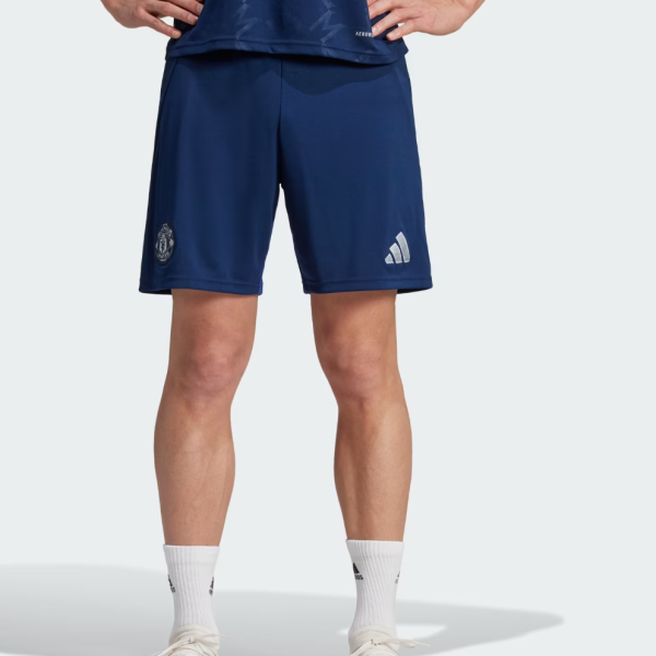 Person shown from the waist down wearing navy blue soccer shorts with a club badge on the left leg and a logo on the right; white crew socks and white cleats are visible, standing with hands on hips.