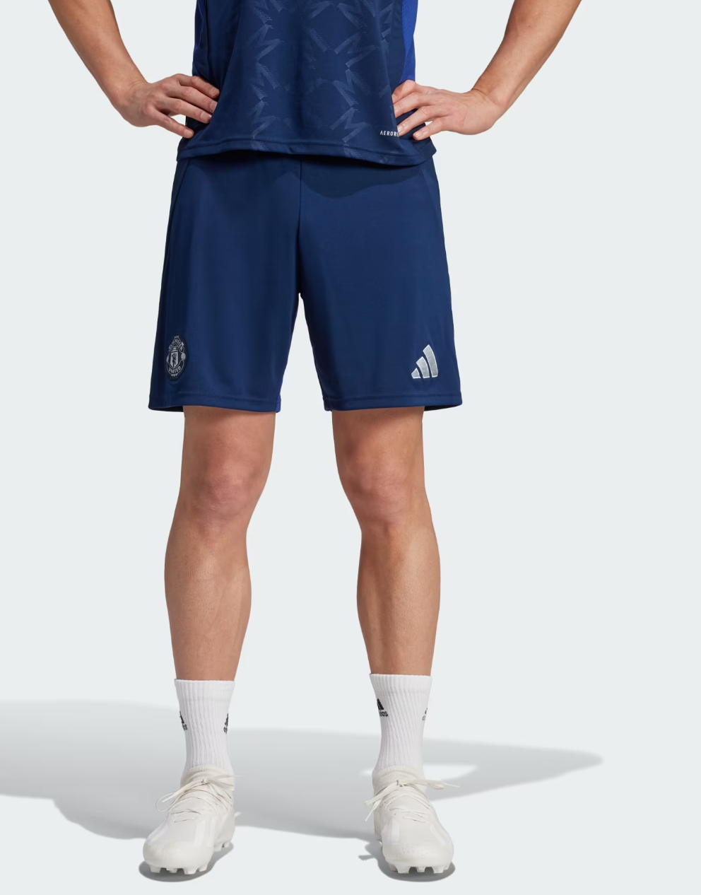 Screenshot_2024-09-18_222513 | UAE SPORT STORE Person shown from the waist down wearing navy blue soccer shorts with a club badge on the left leg and a logo on the right; white crew socks and white cleats are visible, standing with hands on hips.