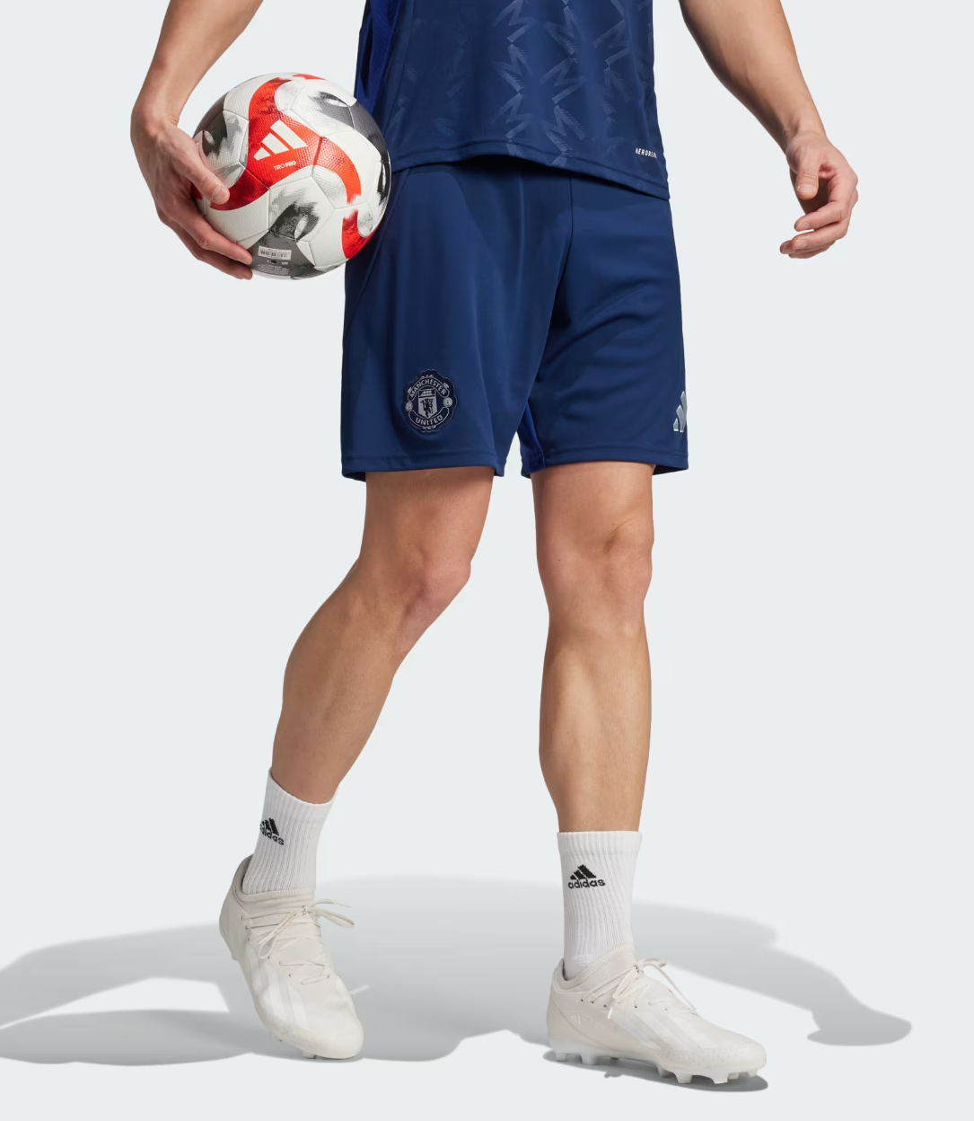 Screenshot_2024-09-18_222533 | UAE SPORT STORE Athlete wearing blue Manchester United shorts holds a soccer ball at his side, wearing white Adidas socks and cleats.