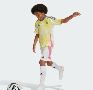 Young soccer player in a yellow Juventus jersey kicks a ball on a light background, mid-stride and smiling.