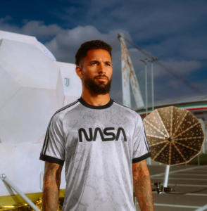 Man with a beard wearing a gray NASA T-shirt stands outdoors near a space-themed sculpture and a large satellite-like dish in the background.