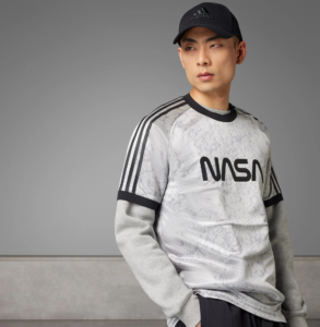 Male model wearing a light gray NASA sweatshirt with black Adidas cap, posing against a gray backdrop, sleeves rolled up.