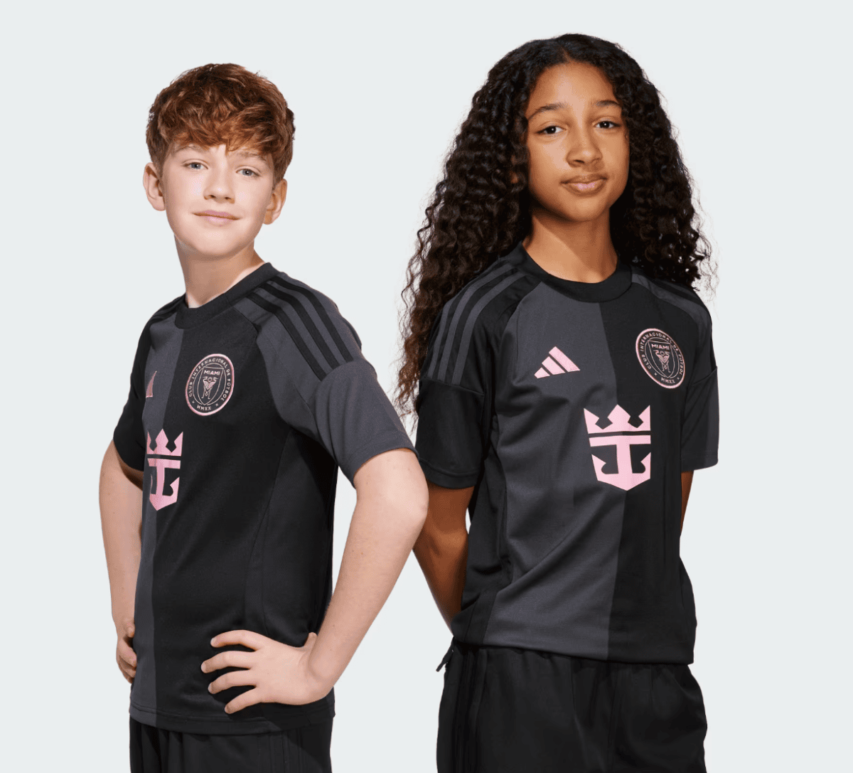 Two children pose in black and gray sports jerseys with pink crest and star-like logo on chest, standing against a light gray background.