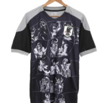 Dark short-sleeve sports jersey with a grayscale anime character grid and a Japan JFA patch, shown on a hanger.