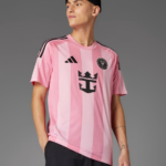 Person wearing a pink striped soccer jersey with a black crest, Adidas logo, and a black beanie against a gray backdrop.