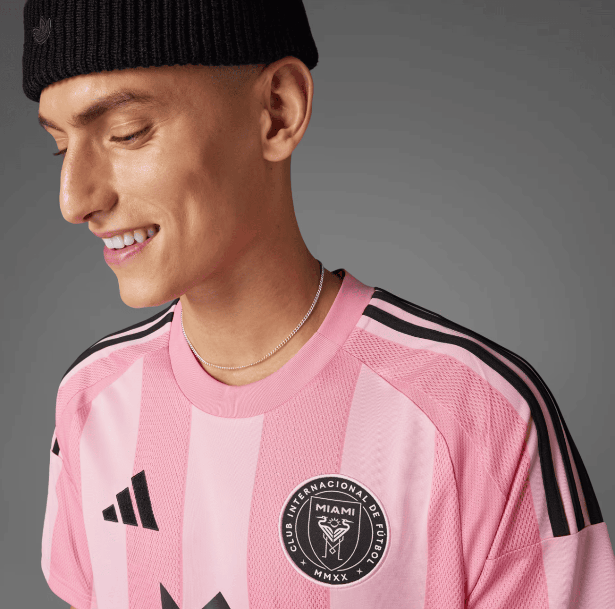 Portrait of a man wearing a pink Inter Miami CF jersey with black stripes, smiling and looking down, black knit cap, gray backdrop.