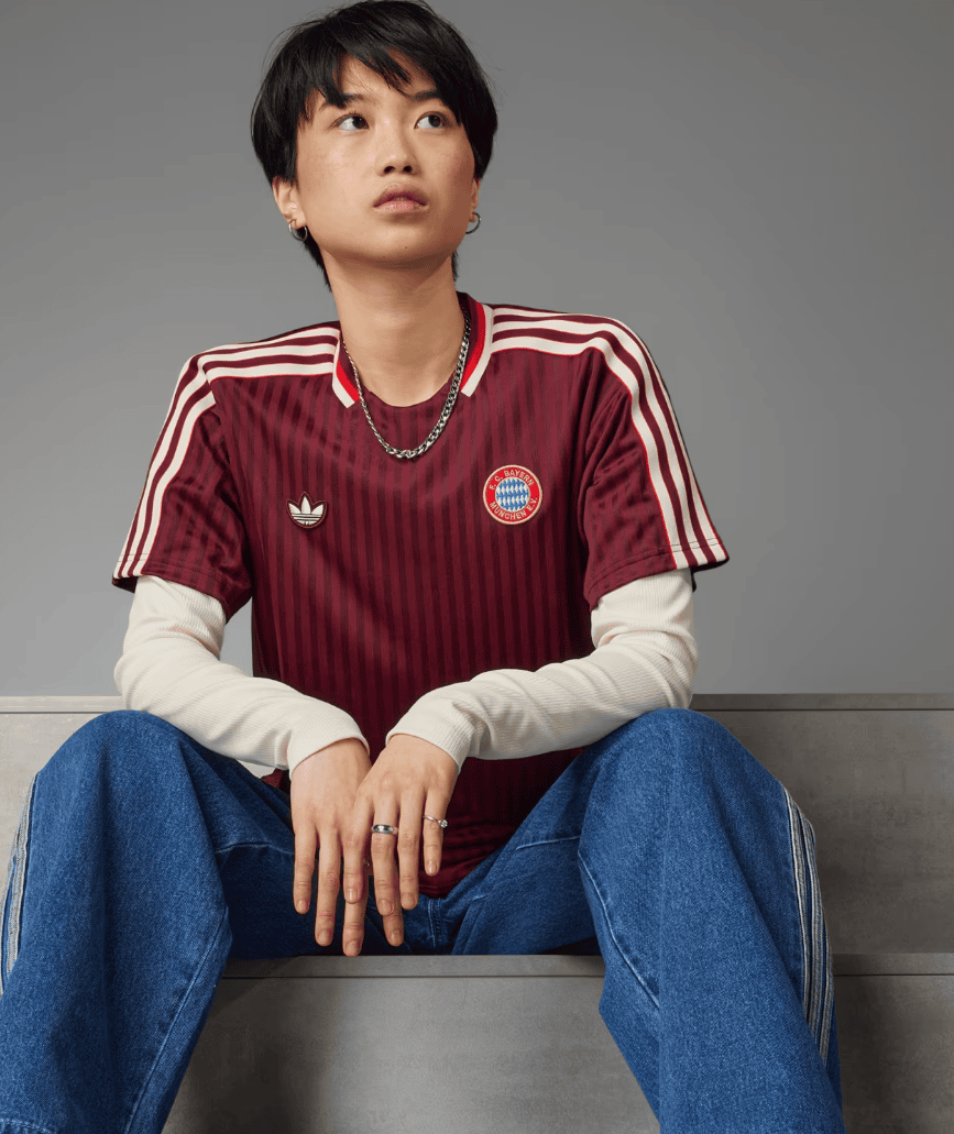 Screenshot_2025-03-21_201153 | UAE SPORT STORE Young person with short black hair wearing a burgundy Bayern Munich jersey and jeans, sitting on a metal bench against a gray background.